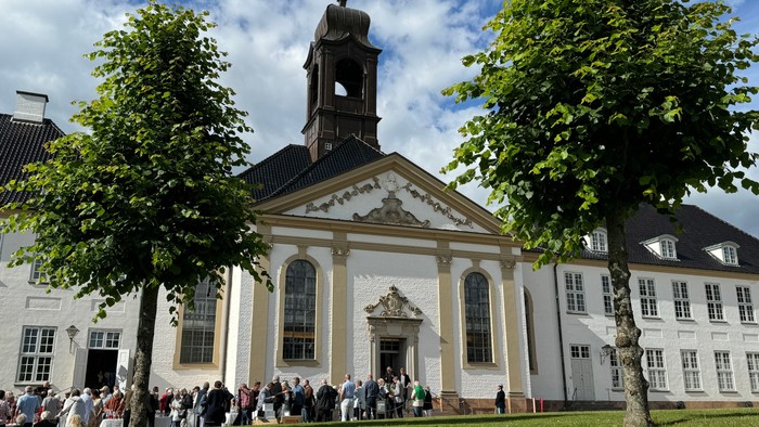 En gruppe af mennesker står foran et stort, hvidt og klassisk bygning med en klokketårn og høje vinduer.