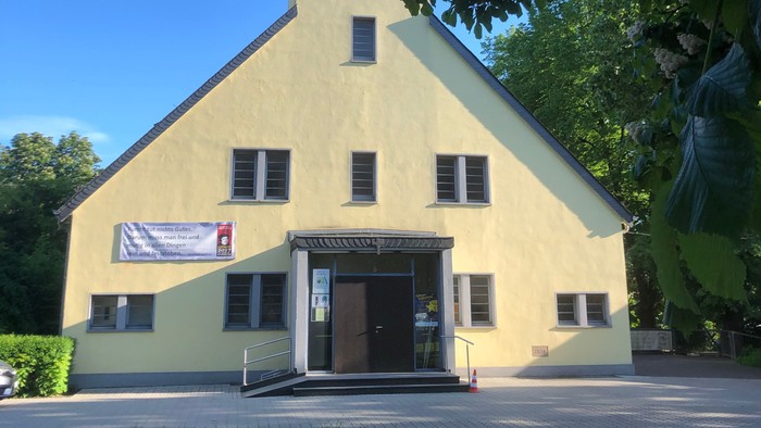 Gelbe Kirche mit Turmspitze und Uhr, Banner über dem Eingang hängend, umgeben von grünen Bäumen und Gras.