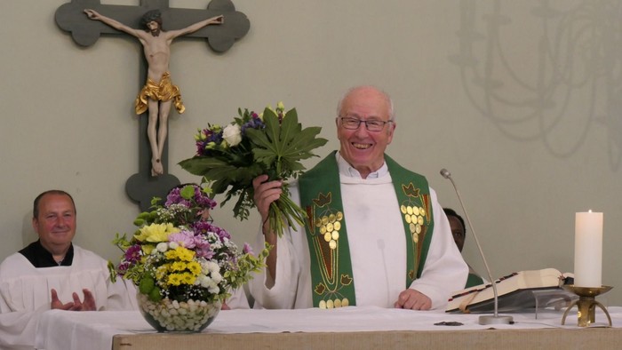 Ein lächelnder Priester in liturgischer Kleidung hält Blumen am Altar einer Kirche während einer Zeremonie.