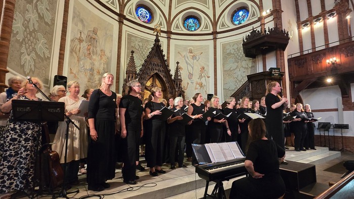 Viele Sängerinnen und Sänger in der Maria-Magdalenen Kirche in Marne, die zum Publikum schauen. Im Vordergrund ist eine Pianisten am Klavier.