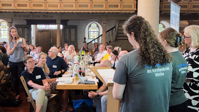 Zwei junge Frauen mit T-Shirts mit der Aufschrift Ev. Jugend im Kirchenkreis Vlotho reden an einem Pult mit einer Frau im Publikum