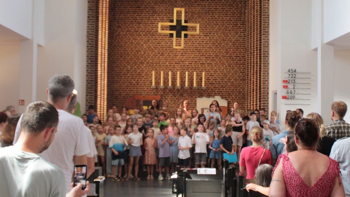 Eine vielfältige Gruppe versammelt sich in einer Kirche zu einem Gottesdienst, wobei Kinder singen und Erwachsene aufmerksam zuhören.