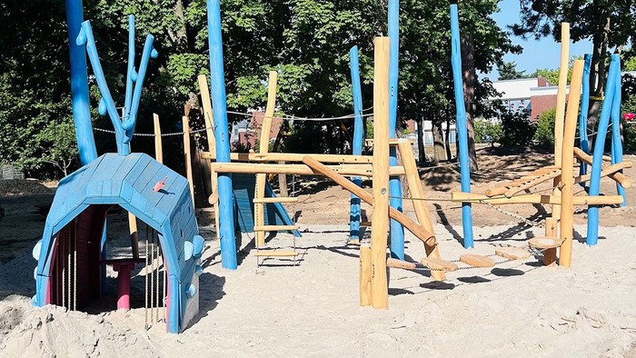 Ein großes blaues Wal-Spielgerät steht als Mittelpunkt eines neu gestalteten Sandspielplatzes mit Kletter-, Balancier- und Hangelelementen aus Holz, der von hohen Bäumen umgeben ist.
