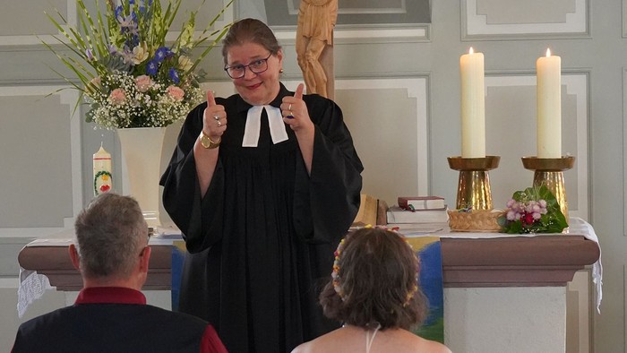 Eine Pfarrerin im Talar steht vor dem Altar einer Kirche und zeigt lächelnd mit beiden Daumen nach oben. Im Vordergrund sitzen zwei Menschen, im Hintergrund sind brennende Kerzen und Blumenschmuck zu sehen.