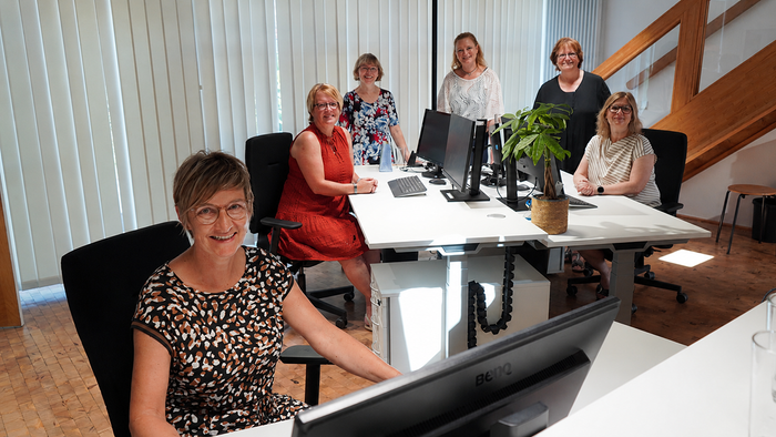Eine Gruppe von fünf Frauen, die in einem gemeinsamen Büroarbeitsplatz mit Computern und Pflanzen lächeln.