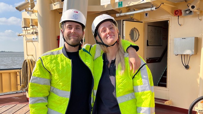 To personer i høj synlighedstøj og hjelme poser på et skibsdæk.