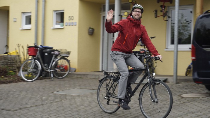Ein Mann in einer roten Jacke fährt mit dem Fahrrad vorbei und winkt, während er an einem gelben Gebäude mit abgestellten Fahrrädern vorbeikommt.