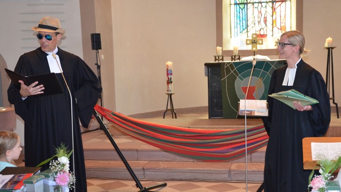 Zwei Geistliche in Roben führen eine Zeremonie in einer Kirche durch, im Hintergrund hängt bunter Stoff.