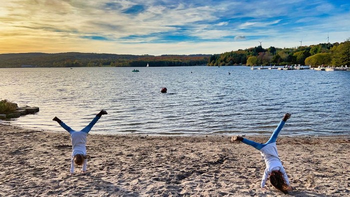 Zwei Kinder schlagen am Strand ein Rad, dahinter ein See.