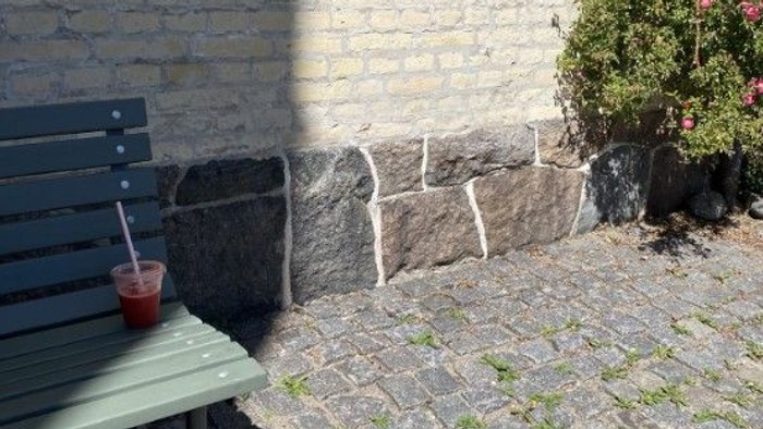 En bænk med en drink står ved siden af en stenvæg og en blomstrende busk på en solbeskinnet terrasse.