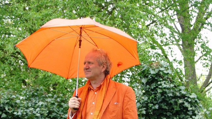 En person i lysende orange påklædning holder en orange paraply udendørs midt i frodig grønne omgivelser.