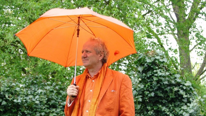 En person i lysende orange påklædning holder en orange paraply udendørs midt i frodig grønne omgivelser.