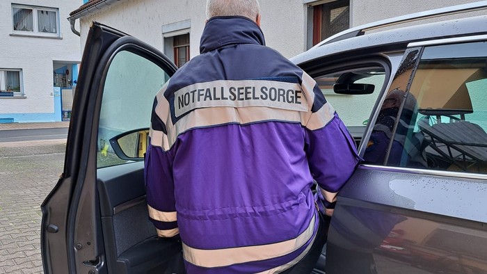 Rettungskraft mit lila Warnweste öffnet eine Autotür mit der Aufschrift „Notfall Seelsorge“.