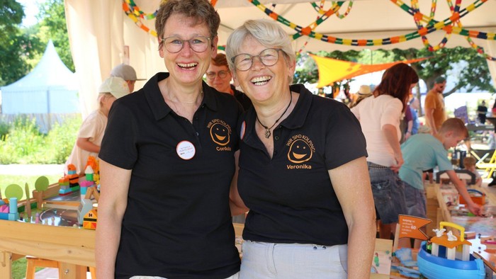 Zwei lächelnde Frauen in passenden schwarzen Shirts posieren fröhlich auf einer bunten Outdoor-Veranstaltung mit einem Spielbereich für Kinder.