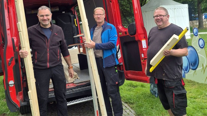 Drei Männer halten große Holzstangen neben einem roten Nutzfahrzeug im Freien.