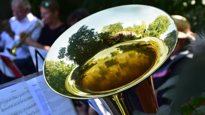 Eine Messingtuba spiegelt Grünpflanzen wider, während ein Musiker im Freien spielt und Notenblätter vor sich hat.