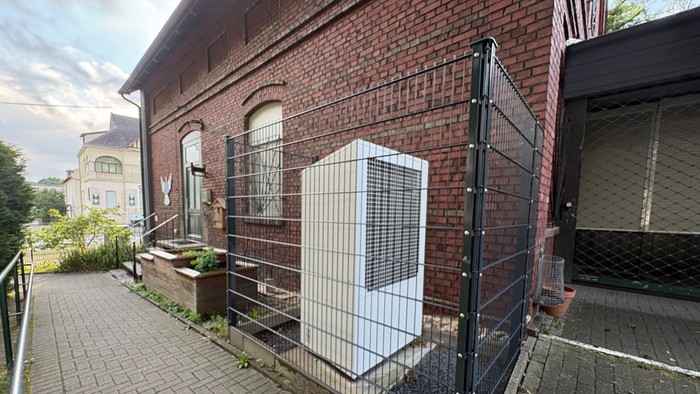 Vor einem Backsteinhaus steht ein großer weißer Kasten mit Lüftungsschlitzen