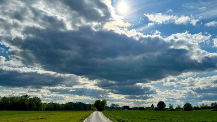 Eine ruhige Landstraße schlängelt sich durch üppige grüne Felder unter einem dramatischen, sonnenbeschienenen Himmel voller Wolken.