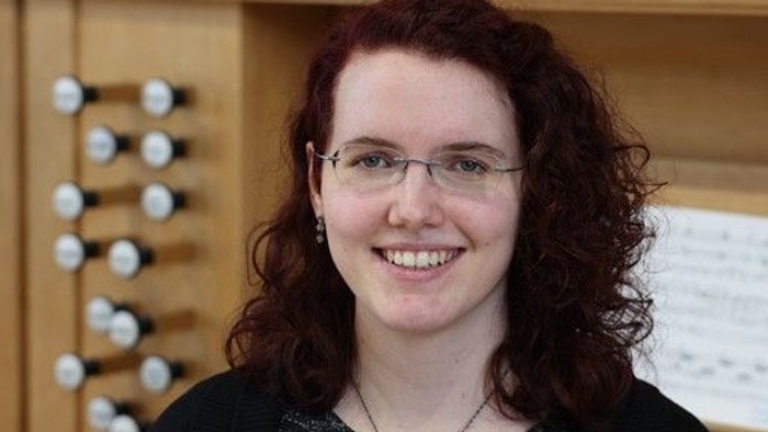 Eine Person mit lockigem Haar und Brille steht lächelnd vor einer Orgel.