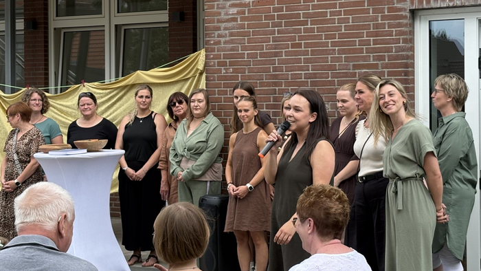 Rund 12 Frauen stehen vor einem Schulgebäude. Sie sind sommerlich und festlich gekleidet. Die Frau vorne im Bild spricht in ein Mikrofon. Menschen hören zu.