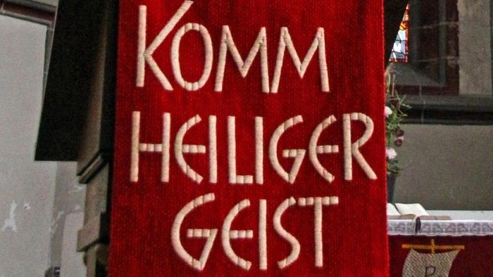Innenansicht einer Kirche mit einem roten Banner mit der Aufschrift „KOMM HEILIGER GEIST“ und bunten Kirchenfenstern.