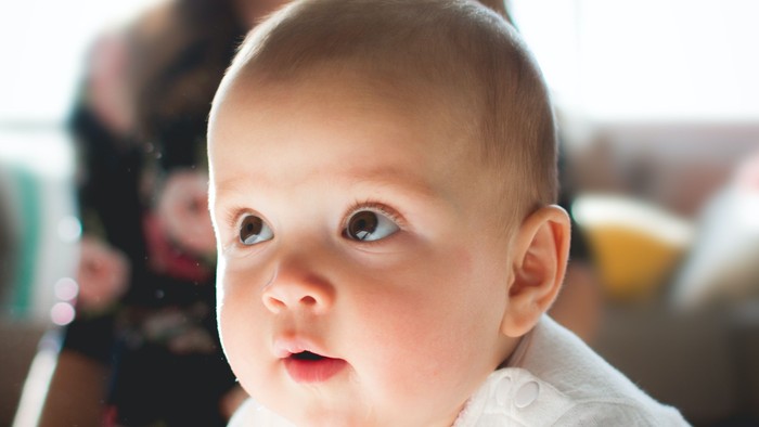 kevin-gent-mVk7pnnlULc-unsplash.jpg Ein neugieriges Baby in einem weißen Kleid schaut nach oben, während ein Erwachsener im Hintergrund lächelt.