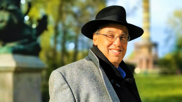 Ein lächelnder Mann mit einem Fedora und einem grauen Mantel steht im Freien in einer parkähnlichen Umgebung.