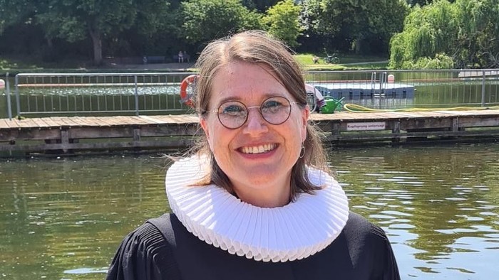 Eine Frau in Abschlusskleidung steht am Flussufer und lächelt.