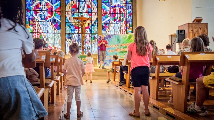 Ein Sänger singt in einem modernen Altarraum, aus dem Mittelgang gesehen. Im Gang stehen mehrere Kinder.