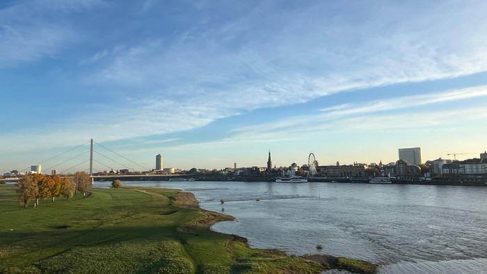 Eine friedvolle Flusslandschaft mit Skyline, grünen grasbewachsenen Ufern und einem klaren blauen Himmel.