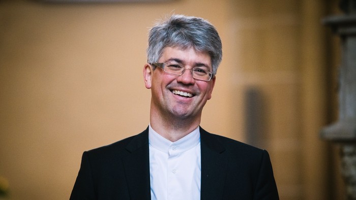 Ein lächelnder Mann in formeller geistlicher Kleidung, der eine Brille und einen schwarzen Anzug mit einem weißen Kollar trägt.