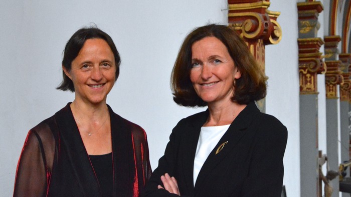 Zwei professionelle Frauen lächeln selbstbewusst in einem eleganten Innenraum mit verzierten Säulen hinter ihnen.