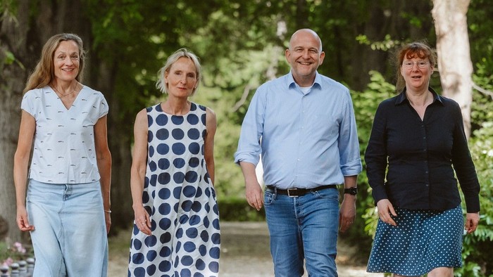 Drei Frauen und ein Mann laufen auf einem Weg unter Bäumen und blicken in die Kamera
