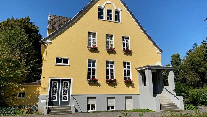 Helles gelbes zweistöckiges Gebäude mit Blumenkästen, eingebettet in eine üppige grüne, sonnige Umgebung.