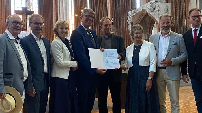 Gruppe von Personen, die in einem festlichen Innenraum mit klassischer Ausstattung offiziell eine Urkunde halten.