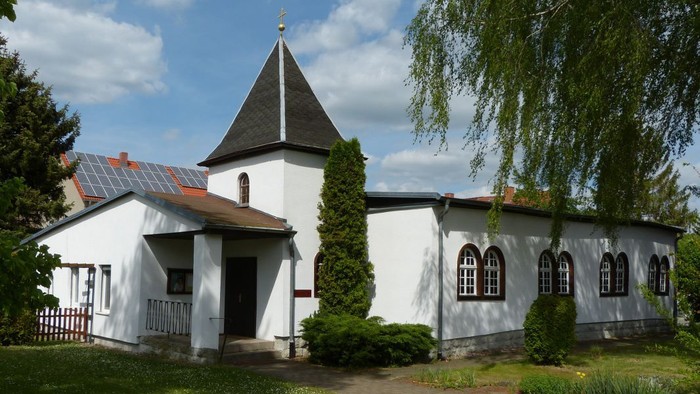 Eine charmante weiße Kirche mit einem steilen Turm und Bogenfenstern, eingebettet in eine üppige, grüne Landschaft.