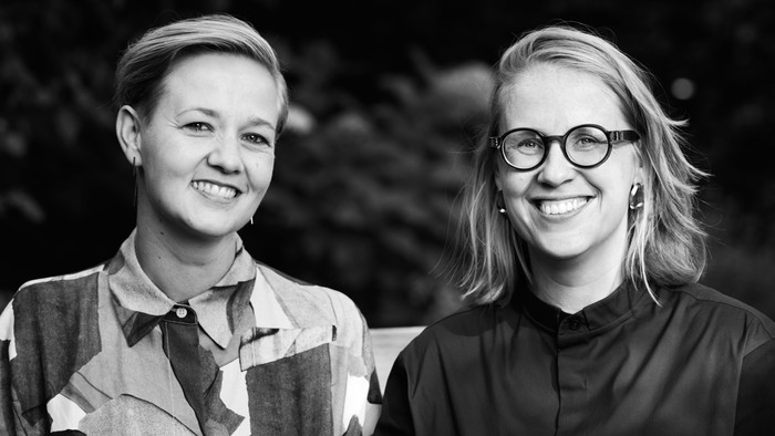 To glade kvinder smiler på et sort-hvidt udendørsportræt med naturlig baggrund.