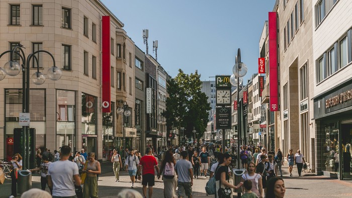 Belebte Stadtstraße mit Geschäften gesäumt, Menschen, die gehen, und hohen Gebäuden unter klarem blauem Himmel.