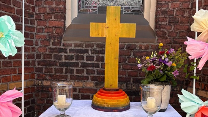 Altar vor alter Kirchenmauer mit einem bunten Sommerblumenstrauß, einem Holzkreuz mit Fuß in Regenbogenfarben, 2 Windlichtern mit brennenden Kerzen