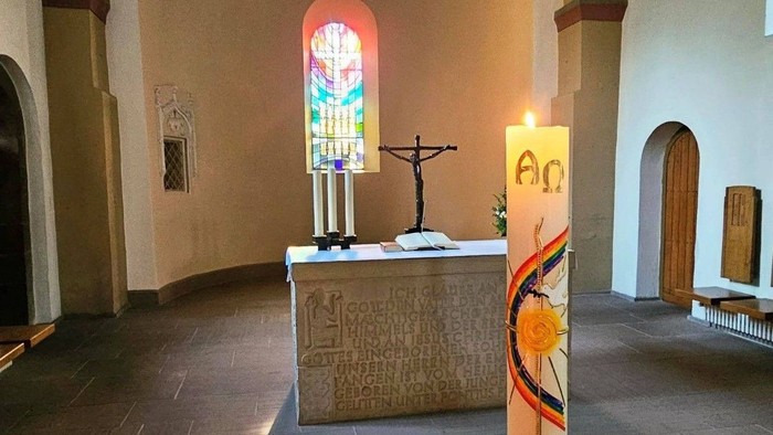 Ein ruhiger Kircheninnenraum mit einem Buntglasfenster, einem Altar mit Kreuz und einer brennenden Kerze.