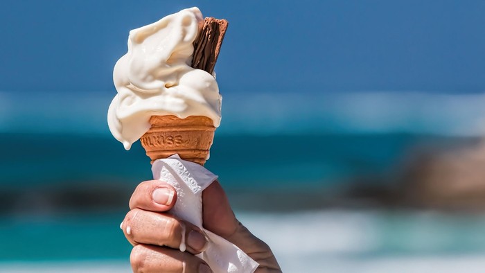Eine Hand hält eine Eiswaffel hoch, Meer ist im Hintergrund.