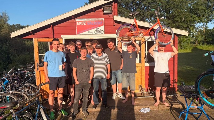 Eine Gruppe meist junger Männer steht vor einer roten Hütte mit vielen Fahrrädern