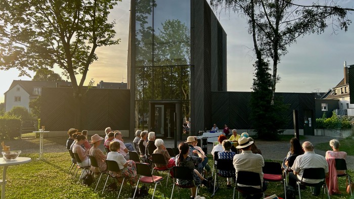 Eine Gruppe von Menschen sitzt auf Stühlen im Freien vor einem modernen Gebäude mit Glasfront.