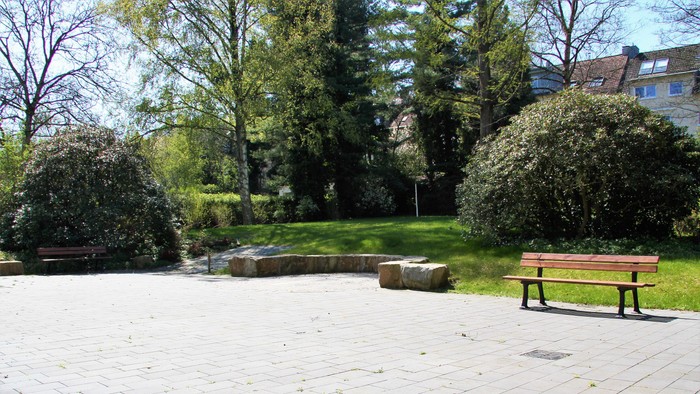 Ein ruhiger Parkbereich mit Bänken, gepflasterten Wegen und üppigem Grün, das ein kleines Wasserspiel umgibt.