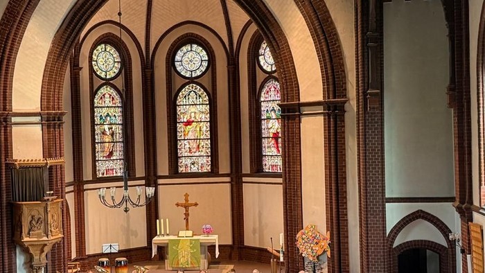 Ein geräumiger Kircheninnenraum mit Bänken, die während eines Gottesdienstes mit Besuchern gefüllt sind, mit bunten Glasfenstern und einem zentralen Altar.