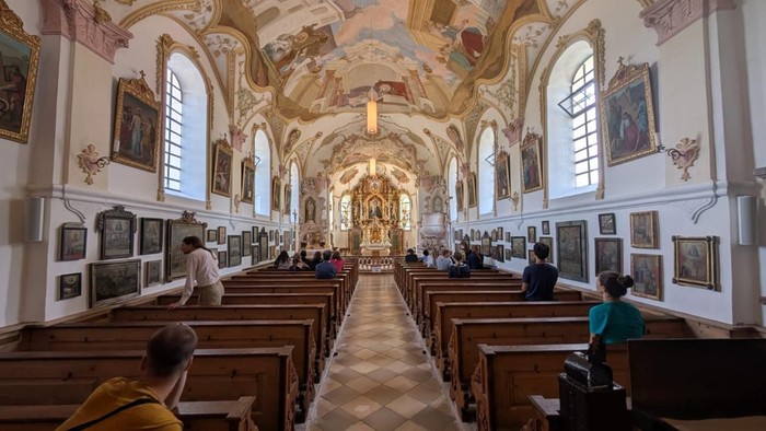 Elegante Kircheninnenausstattung mit prunkvollen Verzierungen, Kirchenbänken und religiöser Kunst an den Wänden während eines Gottesdienstes.