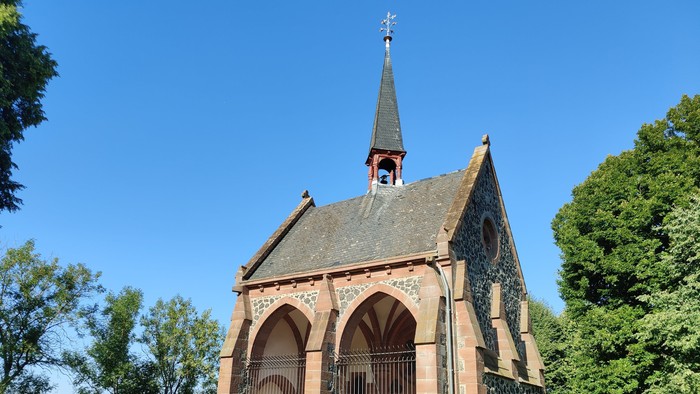Idyllische Kapelle mit gewölbtem Eingang steht in einem ruhigen, gepflegten Friedhof unter klarem blauem Himmel.