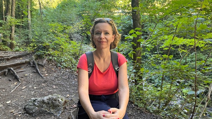 Eine Person sitzt auf einem Waldpfad, lächelt zwischen üppigem Grün und Sonnenlicht, das durch die Bäume fällt.