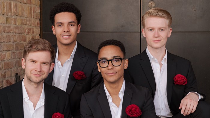 Vier Männer in formeller Kleidung posieren gemeinsam mit roten Rosen-Anstecknadeln vor einem Ziegelwand-Hintergrund.