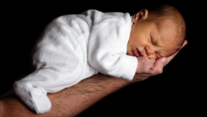 Ein friedliches Baby schläft friedlich auf dem Arm eines Erwachsenen in einem weißen Strampler.